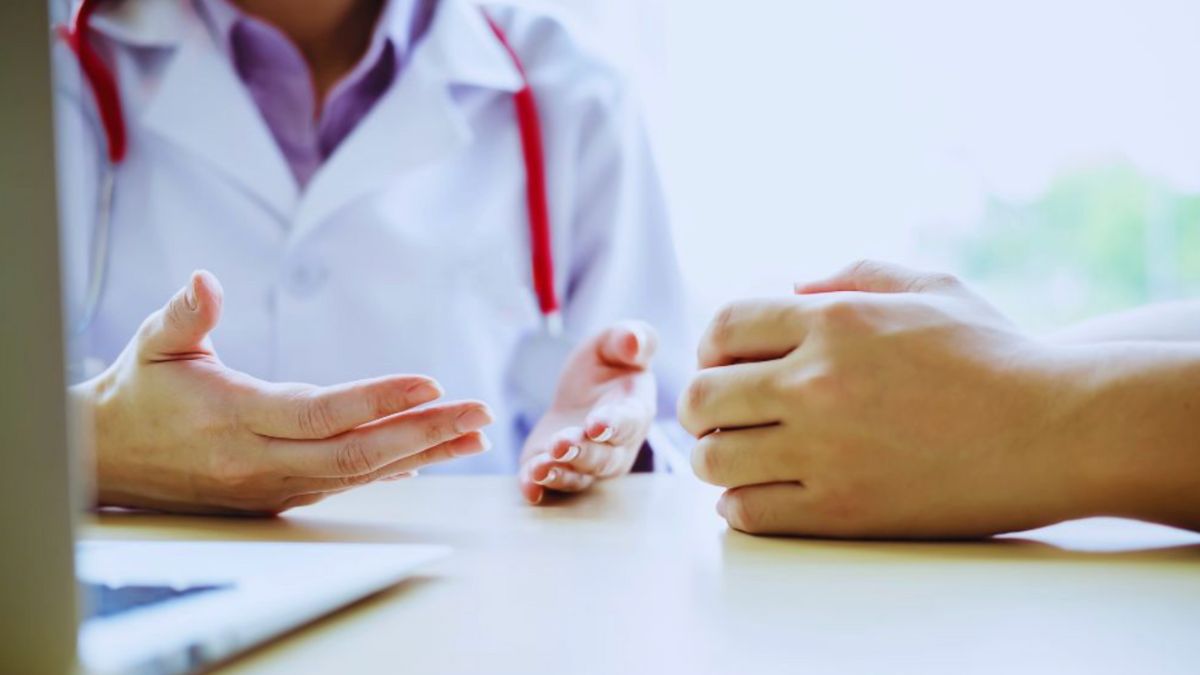 Close-up of a functional medicine doctor in Miami wearing a white coat and red stethoscope, sitting at a desk and consulting with a patient whose hands are visible in the foreground. The scene suggests a personalized, in-depth healthcare discussion in a bright and welcoming medical office.