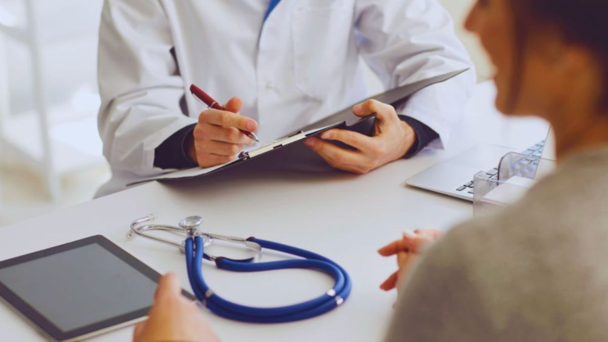 The rheumatologist is in a white coat, speaking with a patient at a desk; the stethoscope and tablet are visible.