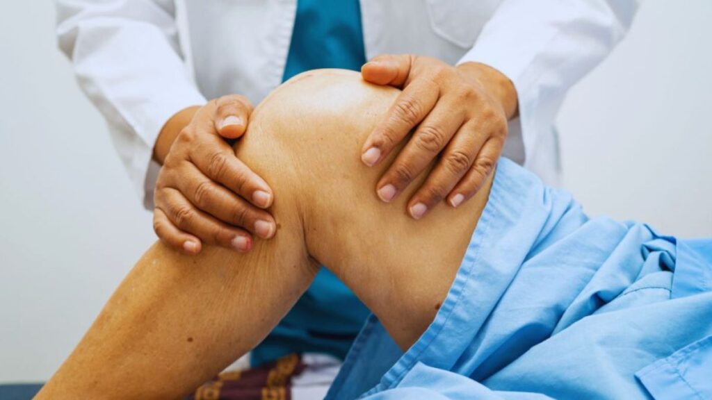 Miami Joint Pain Treatment: How Physical Therapy Can Help 7 A healthcare professional gently examines a patient's knee, applying both hands for assessment or treatment. The patient lies on an exam table wearing blue medical scrubs, representing joint pain treatment in a Miami clinical setting.