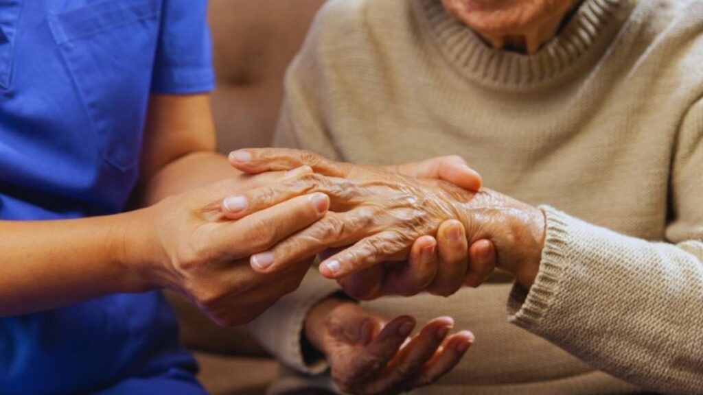 A caring healthcare professional in a blue uniform gently holds and examines the wrinkled hands of an elderly female patient seated on a beige couch. The close-up focus highlights the nurse's supportive grip on the senior's arthritic fingers and palms, conveying compassionate care for joint conditions.